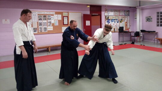 L’image montre trois individus dans un dojo, vêtus de tenues traditionnelles d’arts martiaux. Deux d’entre eux portent un hakama (pantalon large) et semblent exécuter une technique, tandis que le troisième observe. En arrière-plan, un tableau d’affichage contient diverses notes et images, et l’on peut lire l’adresse du site « www.judobruay.jimdo.com ». Des chaises et bancs sont disposés le long des murs.