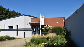 L’image montre un bâtiment moderne avec des murs en briques blanches et rouges. Devant l’entrée, un jardin est aménagé avec diverses plantes et fleurs. L’entrée est constituée de portes vitrées sur lesquelles sont affichées plusieurs notes ou annonces. Le ciel est bleu et dégagé, suggérant une météo clémente.