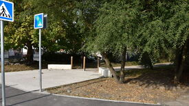 Petite zone pavée entourée d’arbres et de végétation. Deux panneaux de signalisation sont visibles au premier plan : l’un indique un passage piéton, l’autre un chemin partagé pour piétons et cyclistes. À l’arrière-plan, des bâtiments partiellement cachés par la végétation et un sentier qui s’enfonce dans la zone boisée.