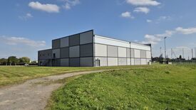 Un grand bâtiment moderne à usage industriel ou logistique est visible, avec une façade grise et des bordures noires. Il est situé dans une zone dégagée avec de l’herbe et un chemin pavé menant à l’entrée. Aucun panneau ou texte n’est visible sur le bâtiment. À l’arrière-plan, on distingue quelques arbres et d’autres structures. Le ciel est dégagé avec quelques nuages épars.
