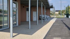 L’image montre un espace extérieur avec un passage couvert soutenu par des poteaux métalliques. Sur la gauche, des portes vitrées donnent accès à un bâtiment dont la façade est brune. Deux portes blanches, chacune munie d’une signalétique, sont visibles sur ce bâtiment. À droite du passage, plusieurs rangées de gradins métalliques sont installées. En arrière-plan, on aperçoit une zone herbeuse et des habitations. Le ciel est dégagé et ensoleillé.