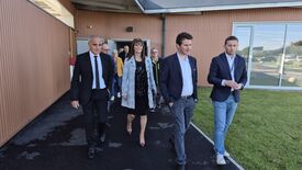 Un groupe de personnes marche à l’extérieur d’un bâtiment aux murs en tôle ondulée brune. Le groupe, composé d’hommes et de femmes en tenue professionnelle (costumes, vestes), semble participer à une visite ou à une conversation. En arrière-plan, une grande fenêtre vitrée reflète l’environnement extérieur, où l’on distingue de l’herbe et des voitures stationnées.