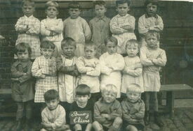 Photographie de groupe montrant des enfants posant pour une photo. Ils sont répartis en trois rangées : la première assise au sol, la deuxième debout, et la troisième debout sur un banc. Ils portent divers vêtements, principalement des chemises à manches longues et des robes. Un petit panneau placé devant le groupe porte l’inscription « Bruay Thiers ».
