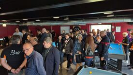 Une salle moderne, aux murs rouges et au plafond avec conduits apparents, est remplie de nombreuses personnes rassemblées autour de plusieurs tables de billard. L’ambiance semble animée, et l’espace est conçu pour accueillir du public.