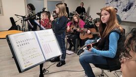 Un groupe de personnes participe à une répétition musicale. Plusieurs jouent de la guitare, une personne chante dans un micro, et une autre joue de la batterie à l’arrière-plan. Au premier plan, un pupitre affiche une partition de la chanson Hey Jude des Beatles, avec les accords, les paroles et des sections surlignées.