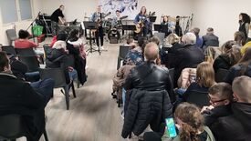 Un groupe de personnes est assis dans une salle, faisant face à une petite scène où un groupe de musiciens se produit. Le groupe comprend plusieurs instrumentistes jouant de la guitare et de la batterie, avec des micros et des pupitres devant eux. Le public, installé sur des chaises, assiste à la performance ; certains regardent leur téléphone. La pièce a des murs clairs et un sol en bois.
