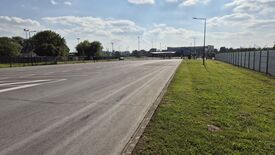 L’image montre un vaste parking vide avec des emplacements de stationnement clairement délimités sur le côté gauche. Sur la droite, une zone herbeuse est bordée par une clôture. À l’arrière-plan, on distingue des arbres et plusieurs bâtiments, dont une structure ressemblant à un poste d’entrée ou de contrôle. Plusieurs lampadaires bordent la route et le parking. Le ciel est partiellement nuageux, laissant passer quelques rayons de soleil qui éclairent la scène.