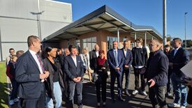 Un groupe de personnes (dont Madame La Maire) se tient à l’extérieur d’un bâtiment. Elles portent des tenues formelles ou semi-formelles, ce qui suggère un rassemblement professionnel ou officiel. Le bâtiment en arrière-plan présente une architecture moderne, avec de grandes fenêtres et un toit en surplomb.
