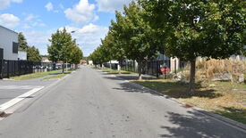 L’image montre une rue droite et vide, bordée d’arbres de chaque côté. Des bâtiments et des clôtures longent la rue, avec un trottoir visible le long de la chaussée. Le ciel est partiellement nuageux. Aucun véhicule ni piéton n’est présent sur la scène.