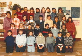Photographie de classe montrant un groupe de 24 enfants et un adulte, probablement un enseignant, posant pour une photo. Les enfants sont répartis en trois rangées : la première à genoux, la deuxième assise sur des chaises, et la troisième debout. L’adulte se tient debout dans la rangée du fond. En arrière-plan, on distingue diverses œuvres artistiques et décorations accrochées au mur.