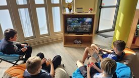 Quatre enfants sont assis sur des poufs et des chaises, jouant à un jeu vidéo sur une télévision. L’écran est posé sur un meuble en bois qui contient également des accessoires de jeu et des décorations, dont une figurine de Pikachu et une Poké Ball. Les enfants tiennent des manettes de jeu et semblent concentrés sur la partie en cours. La pièce est éclairée par la lumière naturelle provenant de grandes fenêtres à panneaux de verre dépoli.