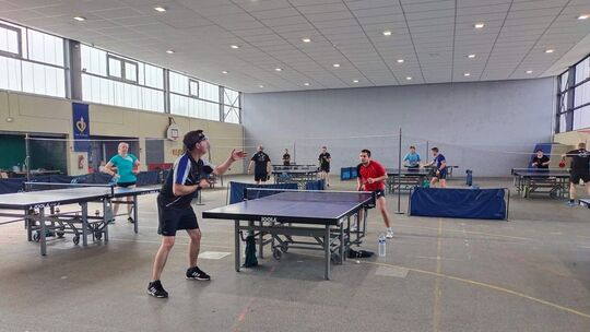L’image montre une salle intérieure dédiée au tennis de table, avec plusieurs tables alignées. Au premier plan, deux joueurs disputent un match : l’un porte un maillot noir et bleu, l’autre un maillot rouge. En arrière-plan, d’autres joueurs sont visibles, soit en train de jouer, soit en attente. La salle est spacieuse, avec un plafond haut, un éclairage suspendu, de grandes fenêtres sur un côté et une bannière accrochée sur le mur gauche.