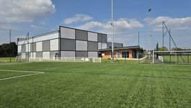 L’image montre un complexe sportif comprenant un grand bâtiment moderne aux façades en panneaux gris et noirs, ainsi qu’une structure attenante en briques avec de grandes fenêtres. À côté du bâtiment se trouve un terrain en gazon synthétique, entouré de filets maintenus par de hauts poteaux et de barrières blanches délimitant le périmètre. Le ciel est partiellement nuageux, laissant passer une lumière claire.
