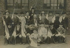 Photographie de groupe prise devant un bâtiment en briques avec de grandes fenêtres. Le groupe est composé d’hommes et de femmes, certains en uniforme, d’autres en tenue civile. Plusieurs personnes tiennent des drapeaux tricolores, suggérant un contexte de célébration ou de commémoration nationale. Aucun texte n’est visible sur l’image.