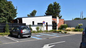 L’image montre un petit parking situé devant un bâtiment moderne aux murs en briques blanches et rouges. Deux places de stationnement réservées aux personnes handicapées sont clairement marquées au sol avec de la peinture bleue et des symboles. Une voiture noire est garée sur une place standard. Un panneau "Stationnement interdit" est visible près de l’entrée du bâtiment, qui est entourée de verdure et de fleurs. Le ciel est bleu et dégagé, suggérant une journée ensoleillée.