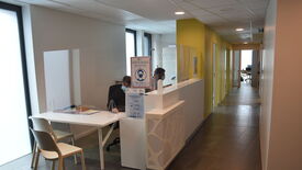 L’image montre une zone d’accueil dans un bureau ou un établissement médical. Deux bureaux sont disposés côte à côte, séparés par une cloison transparente. Une personne est assise à chaque bureau, toutes deux portant un masque facial. Un panneau visible sur le comptoir indique : "PORT DU MASQUE OBLIGATOIRE". L’espace est lumineux, avec des murs blancs et jaunes, et un couloir s’étend à l’arrière-plan, menant à d’autres pièces. Le design est moderne et épuré.