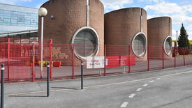 Bâtiment avec trois grandes structures cylindriques en briques, chacune dotée d’une fenêtre circulaire. À l’avant, une clôture métallique rouge porte un panneau indiquant « Collège Jean Macé ». Un autre panneau sur la clôture contient le texte : « Ici, un Espace citoyen sépare l’espace du Département ». L’ensemble semble faire partie d’un établissement scolaire ou d’un équipement public.