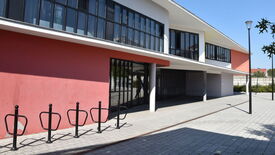 Façade extérieure d’un bâtiment moderne avec de grandes fenêtres et un mur rouge. Plusieurs arceaux métalliques noirs pour vélos sont installés à l’avant. Un lampadaire est visible sur la droite. Le sol est pavé de dalles grises, avec des arbres et des buissons en arrière-plan.
