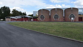 Bâtiment moderne avec trois grandes structures cylindriques à l’avant, chacune dotée d’une fenêtre circulaire. Une clôture rouge entoure la zone d’entrée. Plusieurs bornes blanches bordent l’allée menant au bâtiment. Au premier plan, une route pavée contourne une zone herbeuse. Des arbres sont visibles à l’arrière-plan.