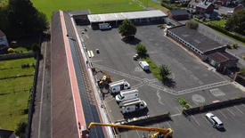 L’image montre une vue aérienne d’un grand bâtiment dont le toit est en cours d’équipement avec des panneaux solaires. Plusieurs véhicules, dont des fourgonnettes et du matériel de chantier, sont stationnés sur une zone pavée adjacente. L’environnement immédiat comprend des champs verts et des bâtiments résidentiels.