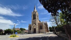 L’image montre une grande église dotée d’un clocher élevé avec une horloge. L’église est située à l’intersection de deux rues. À l’arrière-plan, on aperçoit des arbres et des maisons. Le ciel est dégagé avec quelques nuages. Aucun piéton n’est visible sur l’image. On distingue également des panneaux de signalisation et des poteaux électriques à proximité de l’église.