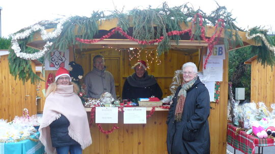 L’image montre un chalet en bois de type marché de Noël, décoré de guirlandes et de guirlandes scintillantes. Quatre personnes sont présentes : deux à l’extérieur et deux derrière le comptoir. Le stand propose divers articles à la vente, dont des peluches et d’autres petits objets. Des tables disposées de chaque côté du chalet présentent également des produits. Des pancartes visibles à l’avant du stand indiquent :

    « BUNSOCKS TELETHON »
    « CAKES APPEAL 2,50 »
