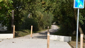 Entrée d’un chemin non goudronné entouré de végétation. Trois poteaux en bois marquent l’entrée. À gauche, un panneau indique « VOIE VERTE DES GUEULES NOIRES ». À droite, un panneau bleu signale un chemin partagé pour piétons et cyclistes. Le sentier s’enfonce dans une zone boisée.
