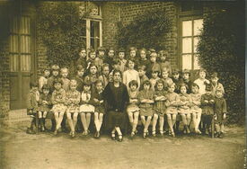 Photographie ancienne représentant un grand groupe d’enfants et un adulte, probablement un enseignant, posant devant un bâtiment en briques avec des fenêtres et de la végétation. Le groupe est organisé en trois rangées : la première avec des enfants assis, la deuxième avec des enfants debout, et la troisième avec des enfants plus grands également debout. L’adulte est assis au centre de la première rangée.