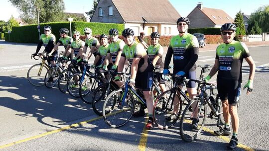L’image montre un groupe de cyclistes debout avec leurs vélos sur une route. Tous portent des maillots de cyclisme verts et noirs assortis, ainsi que des casques. En arrière-plan, on aperçoit des maisons résidentielles, des arbres et un ciel dégagé, suggérant une sortie par temps clair.