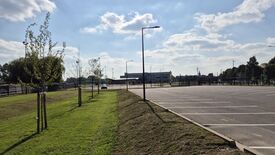 L’image montre un parking vide avec des places de stationnement délimitées sur le côté droit. Sur la gauche, une bande herbeuse est plantée de jeunes arbres, bordée par plusieurs lampadaires. À l’arrière-plan, on aperçoit un bâtiment, probablement une installation sportive ou un bâtiment industriel, ainsi que quelques arbres supplémentaires. Le ciel est partiellement nuageux avec des zones de ciel bleu visibles.
