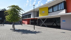Bâtiment moderne avec une façade colorée composée de sections rouges, jaunes et blanches. De grandes fenêtres et un auvent soutenu par des colonnes en forme de X caractérisent l’architecture. Au premier plan, un arbre projette son ombre sur une zone pavée. Un lampadaire est également visible près de l’arbre.