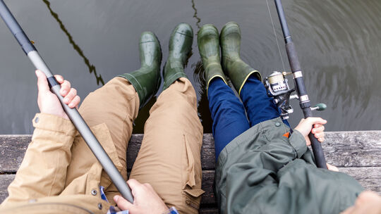 L’image montre deux personnes assises côte à côte sur un ponton en bois, en train de pêcher. Elles portent toutes deux des bottes en caoutchouc vertes. La personne à gauche porte un pantalon beige, tandis que celle à droite est vêtue d’un pantalon bleu et d’une veste verte. Chacune tient une canne à pêche. L’eau en dessous est calme, avec de légères ondulations.