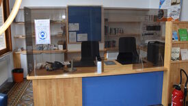 L’image montre un espace d’accueil ou d’information, probablement situé dans une bibliothèque ou un bureau. Le bureau est en bois avec un panneau frontal bleu, et deux chaises noires sont placées derrière. Un écran de protection transparent est installé sur le bureau, accompagné d’un panneau à gauche indiquant "PORT DU MASQUE OBLIGATOIRE" avec une icône de personne masquée. Derrière le bureau, des étagères contiennent des livres et divers objets, ainsi que des panneaux d’affichage avec des documents et annonces. Sur le bureau, on trouve des téléphones, des papiers et un flacon de gel hydroalcoolique. À droite, un extincteur est posé au sol à côté d’autres étagères contenant des livres et des brochures.
