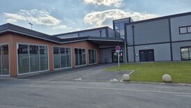 L’image montre un bâtiment moderne avec de grandes fenêtres en verre et une façade mêlant des éléments métalliques et en bois. À l’avant, deux places de stationnement réservées aux personnes à mobilité réduite (PMR) sont clairement identifiables grâce au symbole international d’accessibilité peint au sol. Un panneau de signalisation bleu barré de rouge indique une interdiction de stationner ou de s’arrêter, sauf pour certains véhicules comme précisé par la mention "SAUF". Le ciel est partiellement nuageux et une zone herbeuse est visible sur la droite de l’image.