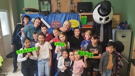 Un groupe d’enfants et d’adultes pose ensemble dans une pièce. Certains enfants tiennent des pancartes vertes avec les mots « SEMAINE », « 2023 » et « BLEUE ». Sur la droite, une grande silhouette vêtue de noir, portant un masque blanc et un haut-de-forme, est visible. En arrière-plan, on distingue divers objets, dont des imprimantes 3D, des étagères et des affiches en français avec les textes suivants :
« Avoir beau jeu de... »
« ...le jouet de l'illusion »
« ...bonne semaine bleue »