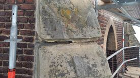 L’image montre une section d’un mur ancien composé de briques et de gros blocs de pierre. Les pierres présentent des signes d’usure et de la mousse est visible à certains endroits. Un « X » rouge est marqué sur le bloc supérieur, probablement pour signaler une réparation ou un remplacement. À gauche, un poteau d’échafaudage métallique porte une étiquette orange avec l’inscription « Layher ». En arrière-plan, on distingue d’autres éléments d’échafaudage et une structure en arc faite de briques.