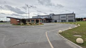 L’image montre un bâtiment contemporain aux teintes brunes et grises, doté de grandes fenêtres et de plusieurs entrées. Devant le bâtiment s’étend un vaste parking, au centre duquel se trouve un espace paysager aménagé avec des plantes et des fleurs. Plusieurs lampadaires entourent la zone de stationnement. Le ciel est couvert de nuages.