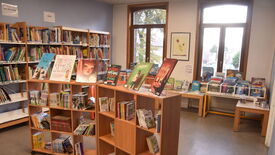 Médiathèque photo 10 Intérieur d’une bibliothèque ou d’une librairie, section jeunesse. Plusieurs étagères en bois sont remplies de livres. Au centre, deux présentoirs mettent en avant des livres pour enfants aux couvertures colorées. En arrière-plan, de grandes fenêtres laissent entrer la lumière naturelle. D’autres étagères et tables avec des livres sont visibles près des fenêtres. Un cadre est accroché au mur, et des panneaux indiquent les sections « DOCUMENTAIRES » et « JEUNES ».