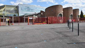 Vue extérieure d’un complexe de bâtiments modernes. À gauche, une façade vitrée encadrée de jaune ; à droite, des tours cylindriques en briques avec de grandes fenêtres circulaires. L’ensemble est entouré d’une clôture métallique rouge. Plusieurs poteaux noirs sont visibles au premier plan. Le ciel est partiellement nuageux.