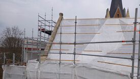 L’image montre un chantier de construction entourant un bâtiment. Des échafaudages en métal, marqués de repères orange, enveloppent la structure. Certaines parties sont recouvertes de bâches blanches, probablement pour des raisons de protection ou de confidentialité. À l’arrière-plan, on distingue un toit haut et pointu, orné d’éléments décoratifs évoquant des flèches ou des tourelles.
