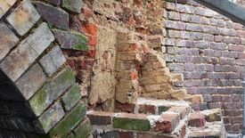 L’image montre un gros plan sur un mur ancien en briques de différentes teintes — rouge, brun et gris — dont certaines sont recouvertes de mousse verte. Le mur présente des signes d’usure, avec des briques manquantes ou endommagées. Deux fils roses sont tendus verticalement sur le côté droit de l’image, probablement utilisés comme repères pour l’alignement de nouvelles briques dans le cadre d’une restauration.