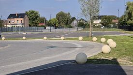 Un parking vide est visible, avec des places de stationnement marquées au sol et une allée courbe au premier plan. De gros plots sphériques en béton sont disposés le long de l’allée et autour d’une zone herbeuse. À l’arrière-plan, on aperçoit des maisons, des arbres et une clôture entourant le parking. Le ciel est dégagé avec quelques nuages.