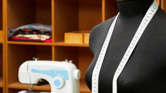 L’image montre un mannequin de couture noir, avec un mètre ruban blanc drapé autour du cou. À l’arrière-plan, on aperçoit une machine à coudre ainsi que des étagères contenant des tissus pliés et des boîtes en bois. L’ensemble évoque un atelier de couture ou un espace de travail textile.