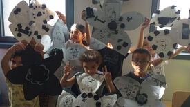 Un groupe d’enfants tient de grandes fleurs en papier décorées de divers dessins. Les visages des enfants sont masqués pour préserver leur anonymat. Les fleurs sont blanches avec des accents noirs, et chaque pétale présente un dessin différent : visages, animaux et autres créations artistiques.