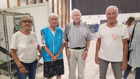 L’image montre quatre personnes debout dans une salle. Deux d’entre elles portent un t-shirt blanc avec un logo en forme de cœur et l’inscription « Les Restos du Cœur ». Une autre personne porte un gilet bleu, et la quatrième est vêtue d’un haut à rayures. En arrière-plan, on aperçoit des cages métalliques et d’autres personnes assises ou debout dans la pièce.
