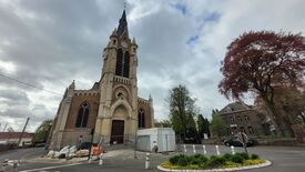 L’image montre une grande église de style gothique, dotée d’un haut clocher avec une horloge. L’architecture présente des arcs en ogive et des détails sculptés. Des travaux de rénovation sont en cours autour de l’entrée, signalés par des barrières et du matériel de chantier. Le ciel est nuageux, et des arbres ainsi que d’autres bâtiments sont visibles en arrière-plan.