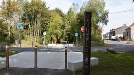 Chemin menant dans une zone boisée, bordé d’arbres et de végétation dense. À l’entrée, plusieurs panneaux sont visibles : un panneau bleu barré indiquant une interdiction d’accès aux piétons et cyclistes, un panneau stop, et un panneau de passage piéton. Au premier plan, un poteau métallique vertical porte l’inscription « VOIE VERTE DES GINETTES NOIRES »