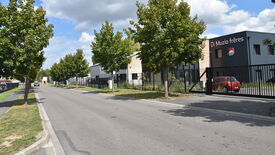 L’image montre une rue bordée d’arbres de chaque côté. Sur la droite, un bâtiment porte l’enseigne "Di Muzio frères" accompagnée d’un logo. Le bâtiment est entouré d’une clôture noire avec un portail, à l’intérieur duquel une voiture rouge est stationnée. Le ciel est partiellement nuageux. Aucun piéton n’est visible sur l’image.