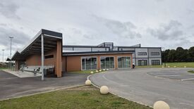 L’image montre un bâtiment moderne dont la façade alterne des murs bruns et gris, avec de grandes fenêtres et portes vitrées. Sur la gauche, une zone couverte est aménagée avec des assises. L’environnement immédiat comprend une route pavée, des places de stationnement, des espaces engazonnés, ainsi que des bornes sphériques en béton disposées le long de la chaussée.