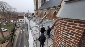 L’image montre un groupe de quatre personnes debout sur un échafaudage installé le long d’un bâtiment en briques, probablement une église ou un édifice similaire. L’échafaudage est équipé de bâches blanches de protection. Le bâtiment présente des fenêtres en arc et un toit incliné recouvert de métal. En arrière-plan, on aperçoit une rue avec des voitures stationnées et des bâtiments résidentiels.